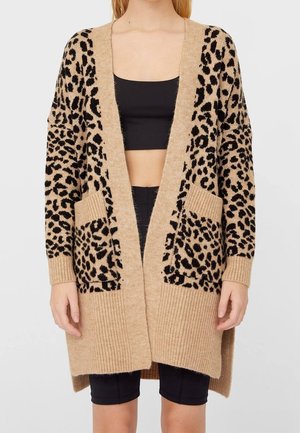 Beżowy długi kardigan w czarne cętki w panterkę, z przednimi kieszeniami, prążkowanym wykończeniem na dole i przy mankietach, noszony otwarty na czarny krótki top i szorty.
