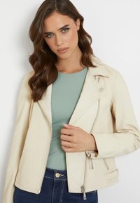 Giacca da biker in pelle crema con zip frontale, colletto diagonale e tasche laterali. Indossata sopra una maglietta verde chiaro e jeans blu scuro.