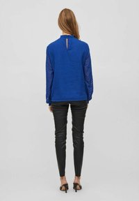 Blue textured top with long semi-sheer sleeves, featuring a keyhole back, paired with black fitted pants and heeled sandals.