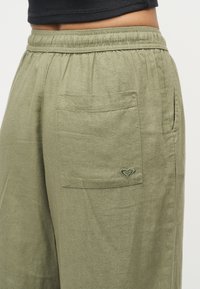 Κοντινό πλάνο χακί παντελονιού με ελαστική μέση, πίσω τσέπη με πάπλωμα και μικρό κεντητό λογότυπο στην τσέπη.