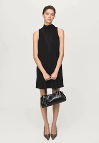 Rochie neagră fără mâneci, cu un gât înalt; modelul ține o geantă mică de mână neagră cu accente aurii; pantofi maro ascuțiți cu un design geometric.