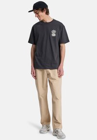 Γκρι βαμβακερό μπλουζάκι με κοντά μανίκια και γραφικό σχέδιο μπροστά. Συνδυασμένο με μπεζ παντελόνι και ανοιχτογκρι αθλητικά παπούτσια. Φοράει απλή καπέλο.