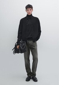 Sort ribstrikket cardigan over en sort rullekrave, kombineret med grå jeans. Tilbehør inkluderer en sort taske og sorte sko. Neutralt baggrund.