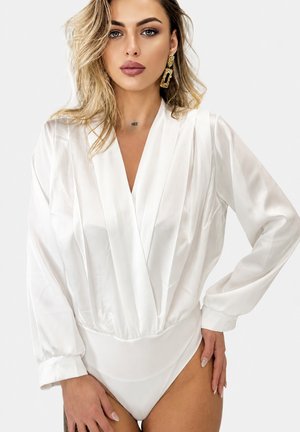 Blondynka z ułożonymi włosami nosi biały jedwabny body z długim rękawem i duże złote kolczyki, pozując z jedną ręką na biodrze.