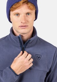 Fleecepullover i mörkblått med en kvartdragkedja. Har en upphöjd logotyp och elastiska ärmslut. Materialet har en mjuk, texturerad yta.