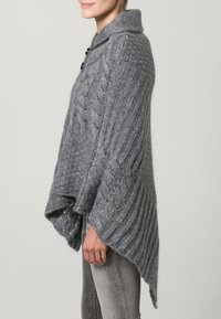 Grå stickad poncho med hög krage, med ett kabelmönster, asymmetrisk design och en knäppning på ena sidan.