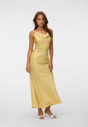 Femme portant une robe maxi en satin jaune sans manches avec un col bénitier et des sandales blanches à talons kitten ouvertes, debout devant un fond uni.