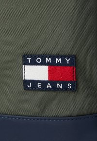 Grön tyg med en texturerad yta har en rektangulär marinblå platta. På plattan står "TOMMY JEANS" i vitt, med en röd, vit och blå logotyp.