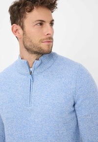 Hellblauer Strickpullover mit Viertelreißverschluss und geripptem Kragen. Weiche Textur, figurbetonte Form und dezentes meliertes Muster. Reißverschluss-Akzent vorne.