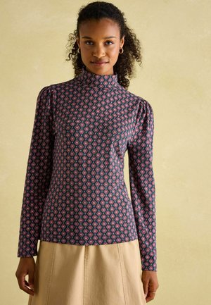 Langärmliges Oberteil aus navyfarbener Stoff mit einem rot-weißen Blumenmuster, hohem Kragen und Puffärmeln, kombiniert mit einem beigen Rock.