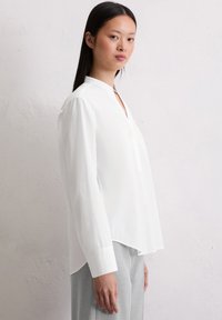 Blusa bianca con colletto, maniche lunghe, una fessura frontale e un orlo posteriore leggermente più lungo. Tessuto liscio, design minimale senza pattern.