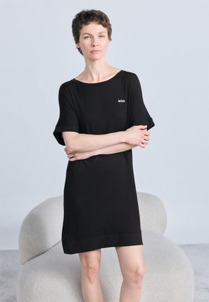 Ženska s kratkimi lasmi, oblečena v črno obleko s kratkimi rokavi z napisom "BOSS", stoji z rokami prekrižanimi pred svetlo sivo ukrivljeno stolom.