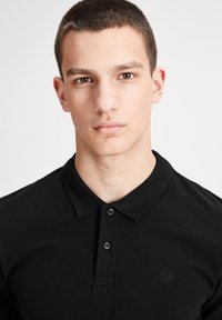 T-shirt polo noir avec un col classique, une patte de boutonnage à deux boutons et un logo subtil sur la poitrine. Tissu lisse avec une coupe ajustée.