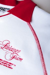 Λευκό πουλόβερ με κόκκινο γιακά και κόκκινες ραφές. Διαθέτει κεντημένη επιγραφή "Athleisure Team" σε κόκκινο, παρουσιάζοντας μια λεία υφή.