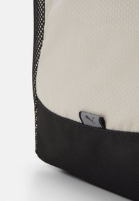 Sac à dos avec une partie supérieure blanche à motifs hexagonaux, une section inférieure en tissu noir, et un petit tag de logo gris sur le côté.
