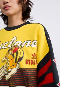 Geel katoenen sweatshirt met fietsgrafiek, zwarte en rode gestreepte accenten en Adidas-logo op de stof gedrukt.