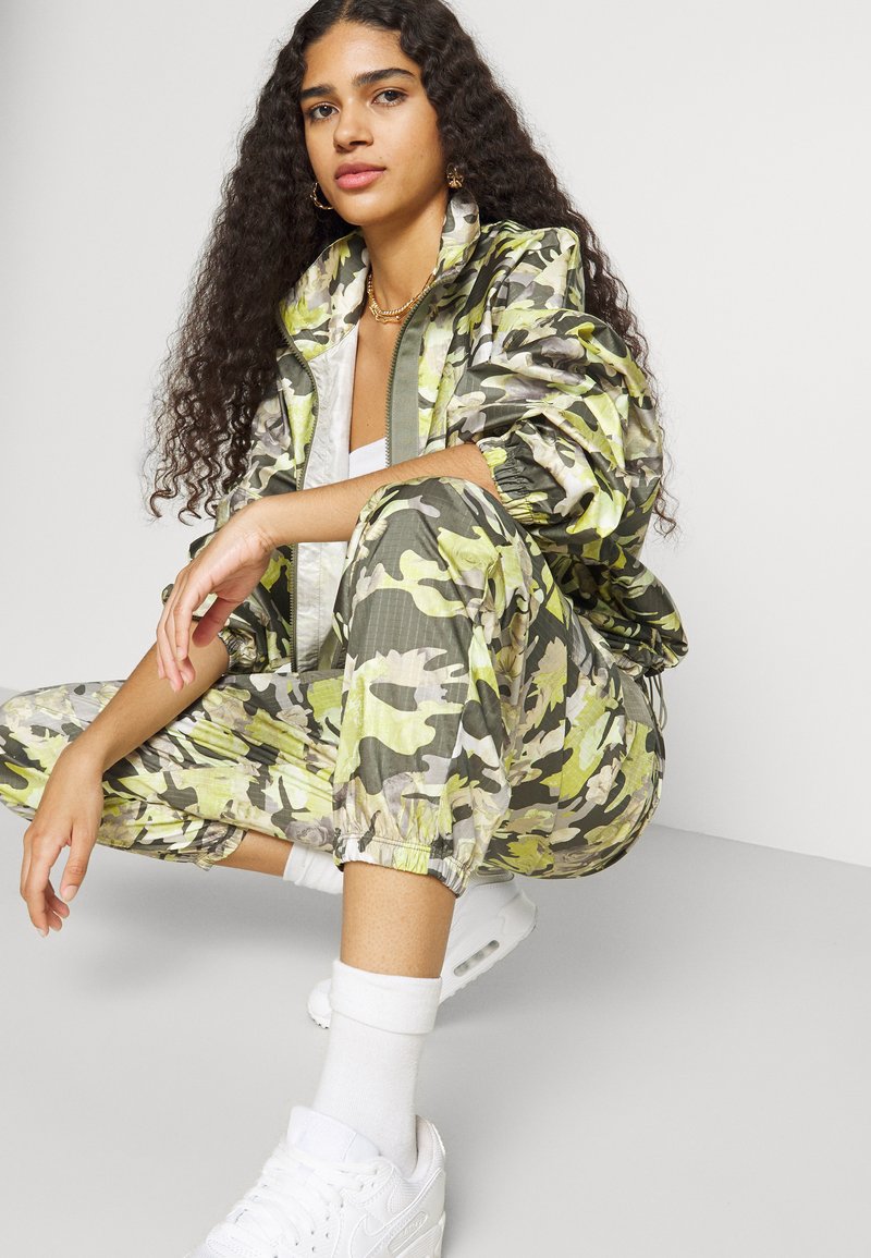 Chándal con patrón de camuflaje en tonos verdes y amarillos, que incluye una chaqueta con cremallera y puños elásticos, combinado con zapatillas blancas y calcetines.