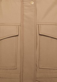 Gros plan sur le devant d'une veste beige avec deux poches à rabat et un bouton-pression sur la patte centrale.