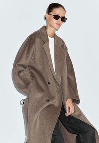Cappotto doppio petto marrone in un tessuto morbido, caratterizzato da maniche ampie e grandi rever, abbinato a occhiali da sole neri e una camicia bianca.