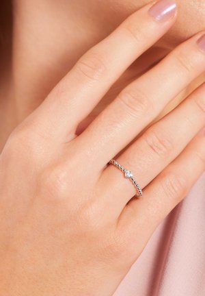 Zilveren ring met een parelvormig ontwerp, met een hartvormige heldere edelsteen. Draag op een hand tegen een zachte roze stof achtergrond.