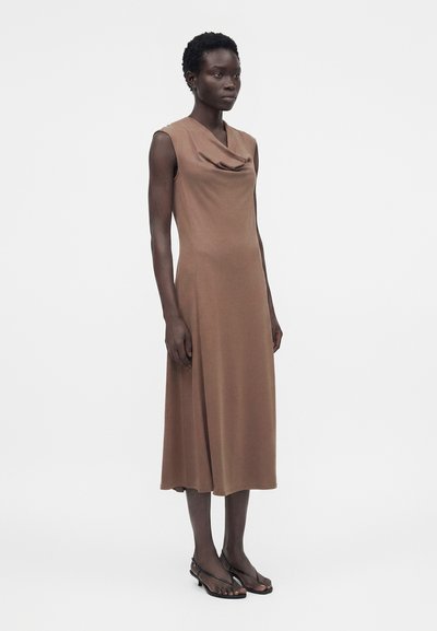 Γυναίκα που στέκεται με καφέ μίντι φόρεμα χωρίς μανίκια και με ντραπαριστό ντεκολτέ, φορώντας μαύρα σανδάλια με τακούνι, σε απλό λευκό φόντο.