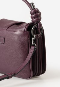 Borsa a mano in pelle viola con superficie texturizzata, manico annodato decorativo, anello in metallo e tracolla regolabile su sfondo bianco.
