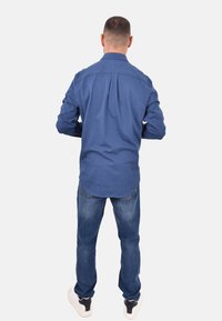 Camicia blu a maniche lunghe con colletto e piega posteriore, indossata con jeans azzurri e sneakers bianche, vista da dietro.