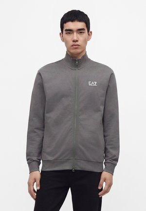 Jeune homme portant une veste zippée grise avec le logo EA7 Emporio Armani sur la poitrine, debout devant un fond blanc uni.