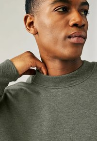 Olivgrön sweatshirt med ribbad krage, åtsittande halsringning och mjuk textur, bärs av en person med en hand på nacken.