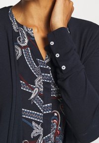 Granatowy kardigan na bluzce z wzorem paisley w odcieniach niebieskiego i bordo. Kardigan ma guziki zdobiące mankiety.