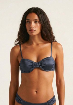 Mujer con cabello largo y rizado que lleva un top de bikini azul oscuro y braguitas a juego sobre un fondo claro y liso.