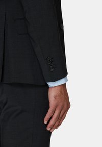 Donkergrijze jas mouw met vier zwarte knopen en een lichtblauwe manchet die onder de jas uitsteekt. Gladde stoftextuur.