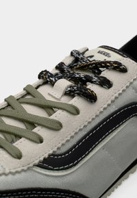 Baskets Vans beige et grises avec une bande latérale noire et blanche, lacets à motif vert et noir noués, vues en gros plan.