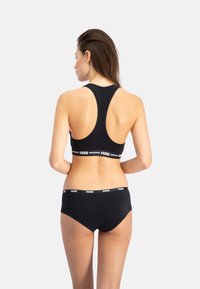 Schwarzer Sport-BH und passende Unterwäsche mit Racerback-Design und elastischem Bund, der das "PUMA"-Logo in Weiß trägt. Glattes, dehnbares Material.