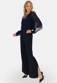 Granatowy kombinezon z przezroczystymi długimi rękawami i dekoltem w serek. Posiada szerokie nogawki z bocznymi rozcięciami. Zestawiony z czarnymi szpilkami w szpic.
