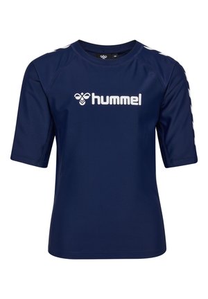 Junge mit lockigem Haar, der ein marineblaues Sportoutfit mit weißen Chevron-Streifen und schwarze Slides trägt und vor einem einfarbigen weißen Hintergrund steht.