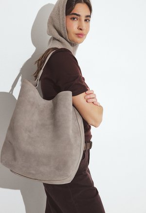 Bolso tote grande de ante gris con una parte superior curva, que cuenta con una sola correa para el hombro y una textura suave, sostenido por una persona que lleva un conjunto marrón.