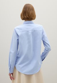 Light blue, long-sleeved shirt with a collar. Made of smooth fabric, featuring a curved hem and buttoned cuffs. Detailed back yoke seam.