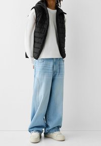 Czarna pikowana kamizelka na białej koszuli z długim rękawem, w połączeniu z jasnoniebieskimi szerokimi dżinsami i kremowymi sneakersami. Proste, codzienne warstwy.