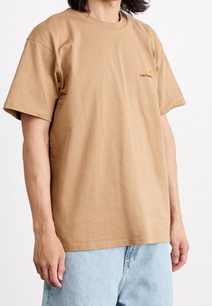 Osoba ubrana w gładki, jasnobrązowy krótki rękaw T-shirt marki Carhartt i jasnoniebieskie dżinsy, stojąca na białym tle.