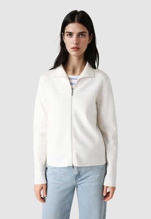 Mujer de cabello oscuro con chaqueta blanca con cremallera sobre una camisa de rayas y jeans azul claro, de pie frente a un fondo gris liso.