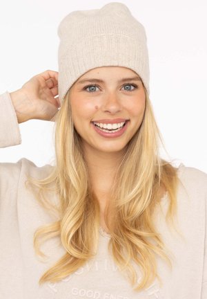 Beige knitted beanie with a ribbed design. Soft texture and round top. Worn by a person with blonde hair, smiling against a plain background.