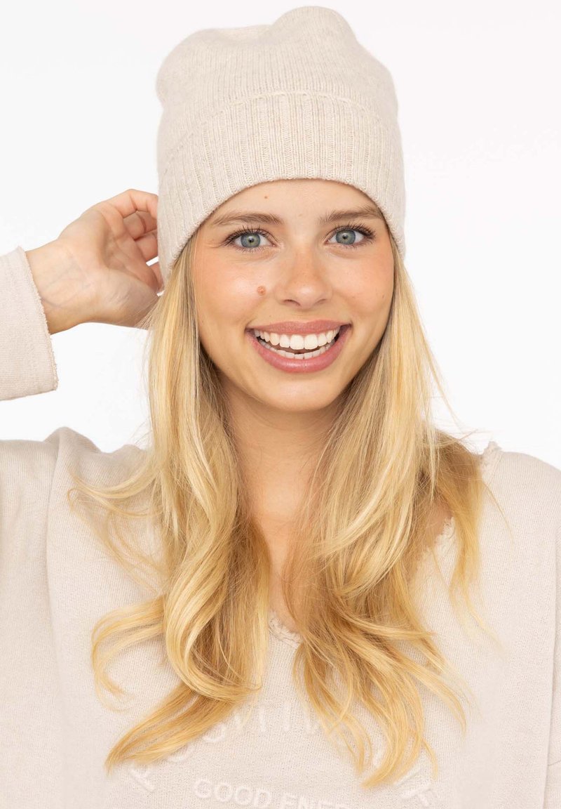 Beige knitted beanie with a ribbed design. Soft texture and round top. Worn by a person with blonde hair, smiling against a plain background.