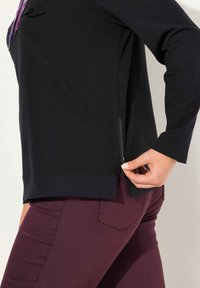 Maglietta nera a maniche lunghe con dettaglio della zip laterale, caratterizzata da una vestibilità rilassata e polsini a coste. Indossata con pantaloni bordeaux.