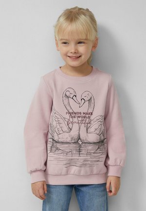 Een jong meisje glimlacht, gekleed in een roze sweatshirt met daarop twee zwanen die een hartvorm vormen, met de tekst "Vrienden maken de wereld mooi."