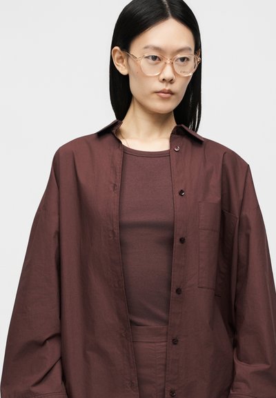 Femme aux longs cheveux noirs portant des lunettes translucides et une chemise ample marron foncé boutonnée par-dessus un haut assorti.