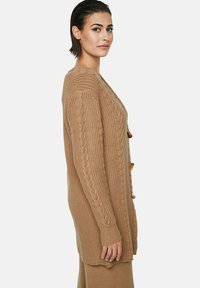 Gilet en maille torsadée beige avec des manches longues, un col en V et des boutons en bois sur le côté. Tissu texturé avec des poignets côtelés.