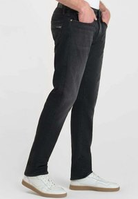 Jean en denim noir à coupe slim, design classique à cinq poches et détails de couture subtils. Portés avec des baskets blanches dotées de semelles beiges.