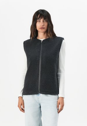 Schwarze Fleece-Weste mit hohem Kragen, Frontreißverschluss und ohne Ärmel. Getragen über einem weißen Langarmshirt, kombiniert mit hellblauen Jeans.