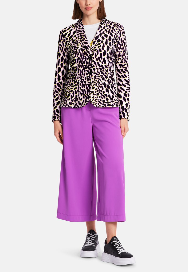 Leopardenmuster-Blazer in Pink, Gelb und Schwarz, kombiniert mit weiten lila Hosen und schwarzen Sneakers mit weißen Sohlen.